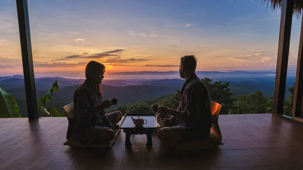 タイ北部の休暇中の男性と女性のカップルは 日の出に木製のバルコニーで紅茶を飲むチェンライドイチャンの山のホームステイキャビン小屋に滞在しています — ストック写真
