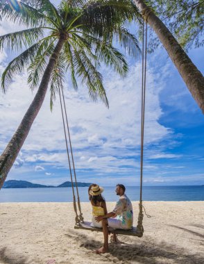Phuket Tayland 'da palmiye ağaçlarıyla sahilde salıncakta sallanan çift. Erkekler ve kadınlar yaz tatili boyunca Tayland 'da tropikal bir plajda salıncakta sallanıyorlar.