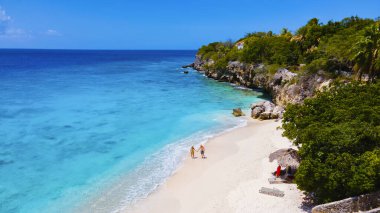 Playa Kalki Plajı Karayip adası Curacao, Playa Kalki Curacao, mavi turkuaz renkli okyanuslu beyaz plaj. Kumsalda yürüyen iki kadın ve erkeğin insansız hava görüntüsü.