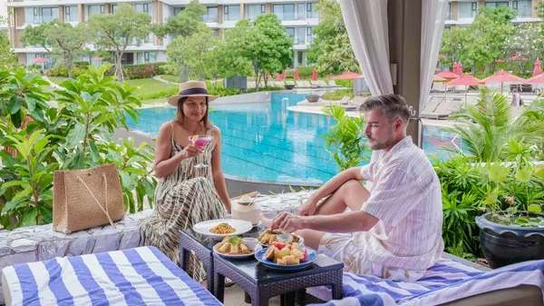 couple having lunch at a restaurant looking out over the swimming pool, a man and woman having dinner in a restaurant by the pool in Pattaya Thailand. Snacks with fruit and spaghetti on a table
