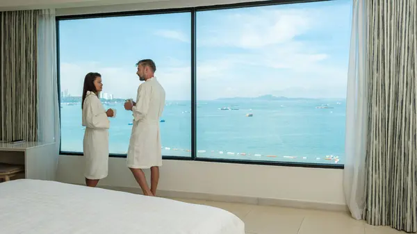 A hotel room with bright fresh colors, minimal style bedroom with ocean view, couple on vacation drinking coffee in the morning looking out the window over the beach and ocean during a luxury holiday