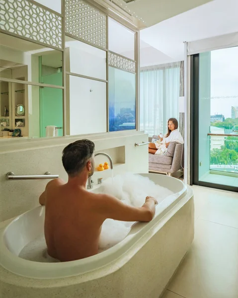 A hotel room with bright fresh colors, a minimal style bedroom with ocean view, men and a bath tub and woman drinking coffee looking out the window at the ocean and beach