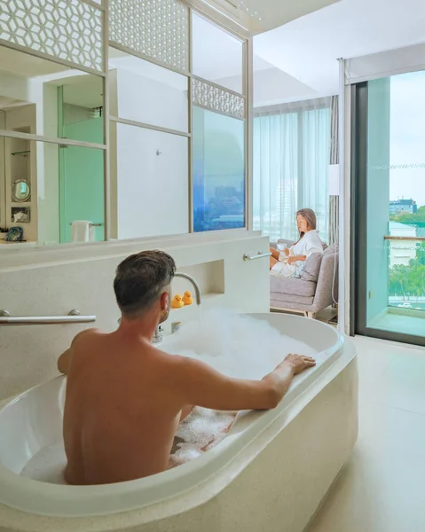 A hotel room with bright fresh colors, a minimal style bedroom with ocean view, men and a bathtub and a woman drinking coffee looking out the window at the ocean and beach