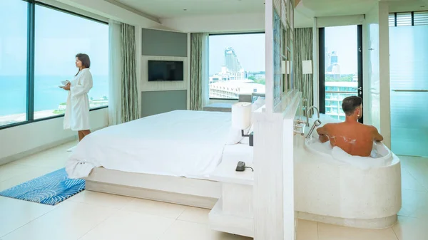 A hotel room with bright fresh colors, a minimal style bedroom with ocean view, men and a bathtub and a woman drinking coffee looking out the window at the ocean and beach