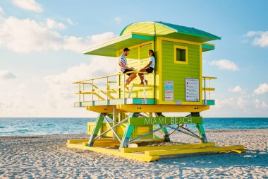 Miami South Beach, Florida US, Miami Beach 'te gündoğumunda cankurtaran kulübesinde bir çift, plajda bir cankurtaran kulübesinde oturan kadın ve erkekler.