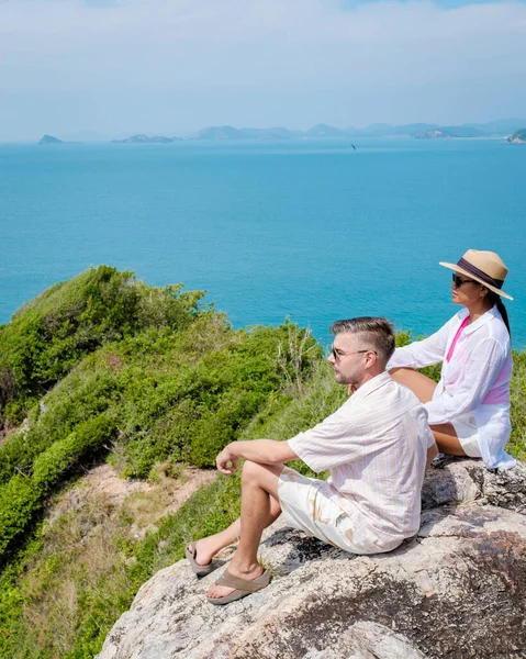 A couple of European men and Asian woman on a boat trip at Ko Kham Island Sattahip Chonburi Samaesan Thailand, a couple sitting on a rock with a view over the ocean at a view point