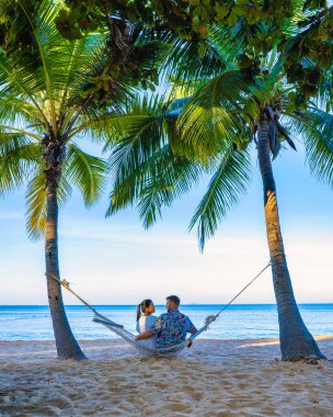 Tropik bir plajda, palmiye ağaçlarıyla gün batımı. Bir çift adam ve orta yaşlı bir kadın, Na Jomtien Pattaya Tayland 'da palmiyeli bir hamakta gün batımını izliyorlar.