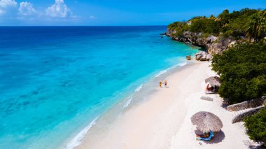 Curacao 'daki Playa Kalki, mavi turkuaz renkli okyanuslu beyaz plaj. Kumsalda turkuaz renkli bir okyanusla yürüyen bir çift kadın ve erkeğin insansız hava görüntüsü.