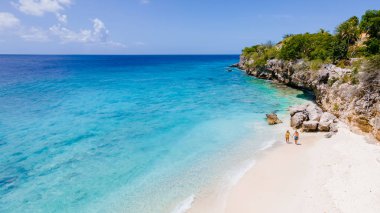 Curacao 'daki Playa Kalki mavi turkuaz renkli okyanusu olan beyaz bir plaj. Curacao 'da bir tatil sırasında sahilde yürüyen bir çift kadın ve erkeğin insansız hava görüntüsü.