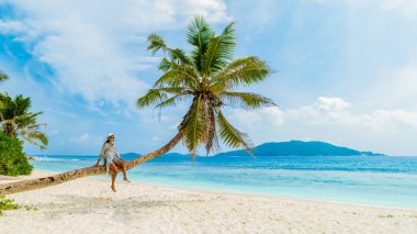 La Digue Seychelles Adaları 'ndaki tropikal bir plajda hindistan cevizi palmiyesinde dinlenen genç bir kadın. Şapkalı Asyalı kadın bir palmiye ağacına oturmuş okyanusu seyrediyor.