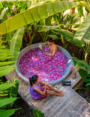 Yaprakları olan yeşil bir bahçede, açık havada bir küvette iki erkek ve bir kadın gün batımında bir banyoda çiçeklerle küvette.