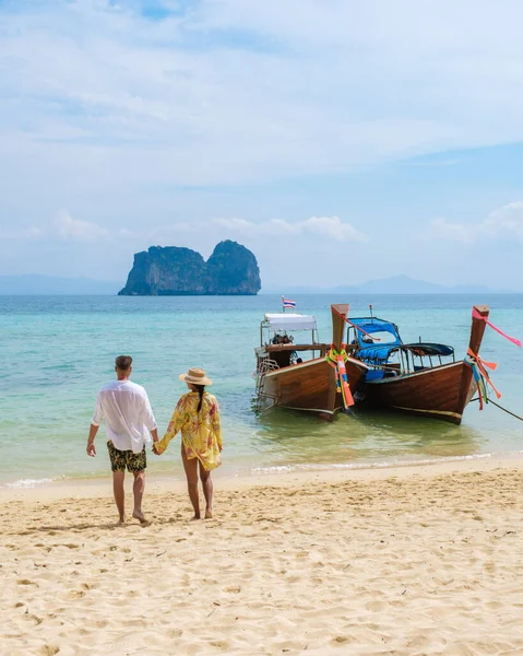 Happy young couple, an Asian woman and European men on the beach of Koh Ngai island, soft white sand, and a turqouse colored ocean in Koh Ngai Trang Thailand with longtail boats on the background