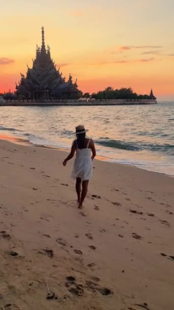 パタヤチョンブリタイのビーチで夕暮れにパタヤのビーチを歩くアジアの女性 真実のサンクチュアリ パタヤタイ 太陽の下で海による木造寺院 — ストック動画