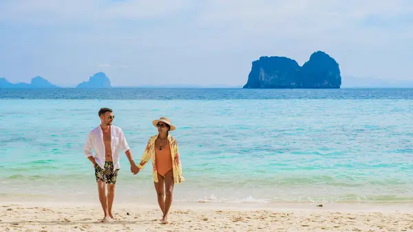 Happy young couple, an Asian woman and European man on the beach of Koh Ngai island, soft white sand, and a turqouse colored ocean on a sunny day, man in swim short and white shirt, woman in bikini