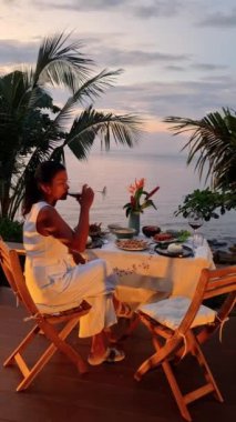 Bulutsuz bir gökyüzünün altında, bir kadın kumsalda bir masada oturmuş, şarabını yudumluyor. Arecales rüzgarda sallanıyor, Koh Chang, Tayland 'da huzurlu bir açık hava ortamı yaratıyor.