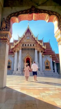 Bir çift görkemli bir tapınağın önünde el ele durur, binanın karmaşık cephesine hayran kalırlar. Simetri mimarisi romantik anı geliştirir Wat Benchamabophit Tapınağı, Bangkok, Tayland