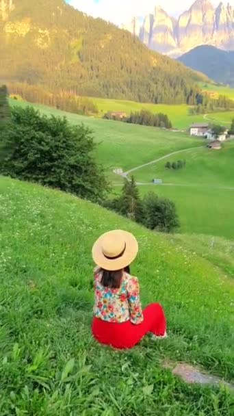 一个戴着太阳帽的女人悠闲地坐在长满青草的小山上 欣赏周围的青山和自然景观 四周环绕着树木和草原 — 图库视频影像