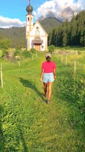 一个穿着短裤的女人正在穿过长满青草的高地走向一座迷人的教堂 绿树成荫的自然景观和宁静的氛围 意大利Val Funes Santa Magdalena — 图库视频影像