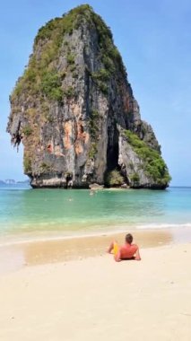 Arka planda yüksek bir kaya oluşumu olan bir adam kumlu sahilde dinleniyor. Doğal manzara, ağaçlar, sakin bir su kütlesi ve Railay Sahili, Krabi, Tayland üzerinde açık bir gökyüzü içerir.