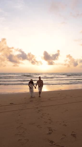 カップルは夕暮れの砂浜に沿って散歩し 手を携えてカラフルな空を眺め 太陽が地平線に沈むように静かな海水を眺めています プーケット — ストック動画
