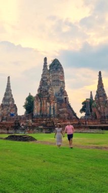 İki kişi yemyeşil bir tarlada antik bir tapınağın arka planında geziniyor. Sitenin ortaçağ mimarisi ve tarihi önemi, turistler için bir hardal oluşturuyor: Ayutthaya, Tayland