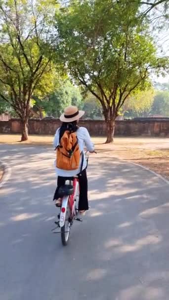 女性は道に沿って自転車をペダルで歩いていて 自転車の車輪がタイヤを回っている 彼女は帽子をかぶっている 木や植物を通過 タイのスクタイ — ストック動画