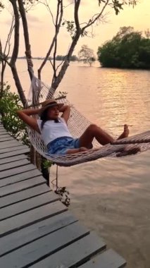Taylandlı Asyalı kadınlar Chantaburi Tayland 'da bir mangrov ormanıyla çevrili nehir kenarındaki bir hamakta uzanıyorlar.