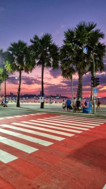 Pattaya Thailand 19 December 2024, A city street lined with towering palm trees, with a crosswalk in the foreground where pedestrians cross. clipart