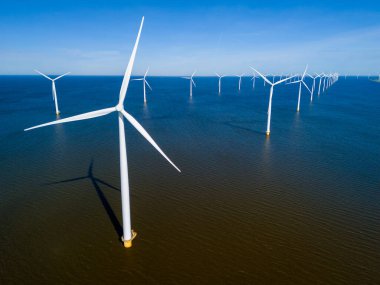 Bir grup yel değirmeni, canlı bahar mevsiminde Hollanda Flevoland bölgesinde zarifçe yüzüyor. Yel değirmeni türbinlerinin insansız hava görüntüsü mavi okyanustaki yeşil enerjiyi gösteriyor