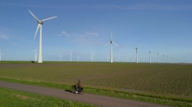 Hollanda 'da bir yel değirmeni çiftliğinin yanında yeşil bisiklet süren genç bir adam Fleordoostpolder, yeşil elektrikli bisiklet ile kırsal alanda bisiklet süren bir adam, yel değirmeni türbinleri çevre dostu yeşil enerji