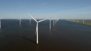 Okyanustaki yel değirmeni parkı, baharda güneşli bir günde Hollanda 'da yeşil enerji üreten yel değirmeni türbinlerinin insansız hava görüntüsü. Mavi okyanus ve mavi gökyüzü
