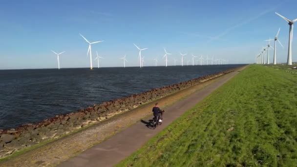 オランダの美しい春の日に風力発電所の風車タービンで電気グリーン自転車の自転車に乗った若い男 ノルドストポルダー 男性自転車の田園地帯の湖畔 — ストック動画