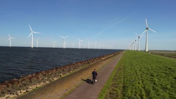在荷兰弗列佛兰的一个风车农场旁边 一个骑电动绿色自行车的年轻人骑自行车 在荷兰的一个堤坝上 男人骑自行车在一个叫Ijsselmeer的湖边 — 图库视频影像