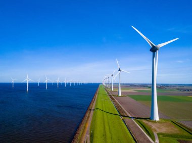 Hollanda, Flevoland 'da, canlı bahar mevsiminde sakin bir su kütlesinin yanında duran bir sıra rüzgâr türbininin sakin bir görüntüsü. Okyanustaki yeşil enerji