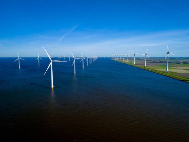 Yel değirmenleri, büyük bir su kütlesinin üzerinde dimdik durur, rüzgarda zarif bir şekilde dönen bıçakları, Hollanda Flevoland 'da bahar mevsiminde yeşil enerji üretiminin büyüleyici bir görüntüsünü yaratır..