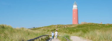 Texel, Hollanda 'da deniz fenerinin yanındaki patikada iki kişi yürüyor. Arka planda dalgalar kıyıya vuruyor. Teksas Hollanda 'nın ikonik kırmızı deniz fenerinde bir çift.