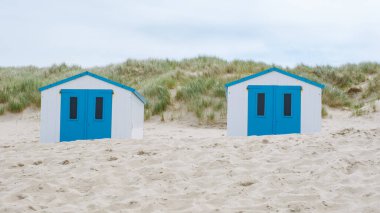 Two colorful beach huts with bright blue doors stand out against the sandy shore, creating a picturesque scene on the coast of Texel, Netherlands. clipart