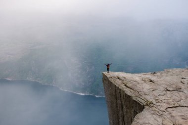 Bir yürüyüşçü, kolları açık bir uçurumun kenarında durur ve sisle kaplanmış Norveç fiyortlarının nefes kesici manzarasını seyreder. Norveç, Preikestolen