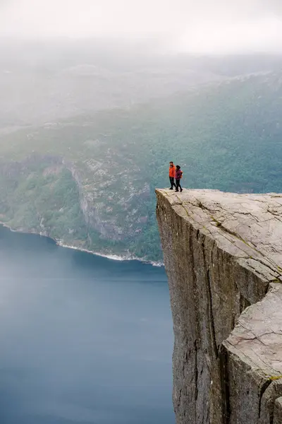 stock image Preikestolen, Norway, Two people stand on the edge of a cliff overlooking a fjord in Norway. The cliff is known as Preikestolen, or Pulpit Rock, and is a popular tourist destination.