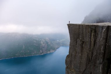 Preikestolen 'in kıyısında tek bir kişi duruyor, Norveç' in dramatik bir uçurumu, sisle kaplanmış engin bir su açıklığına bakıyor. Norveç, Preikestolen