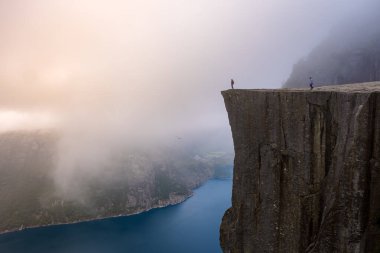 Norveç 'te sisli bir sabah sahnesi. Preikestolen 'in kenarında iki kişi duruyor, ünlü bir uçurum yüzü, bir vadi ve derin mavi bir fiyort manzaralı. Norveç, Preikestolen 'de yürüyüş yapan erkekler ve kadınlar