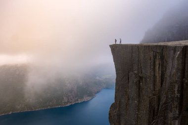 Preikestolen 'in nefes kesen manzarası, Norveç' te uçurum, kenarında iki kişi var. Manzara sis ve sisle kaplı. Dramatik ve hayranlık uyandıran atmosfere yeni bir hava katıyor..