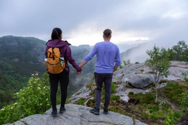 Sisli bir Norveç vadisine bakan kayalık bir çıkıntıda el ele duran bir çift. Norveç, Preikestolen