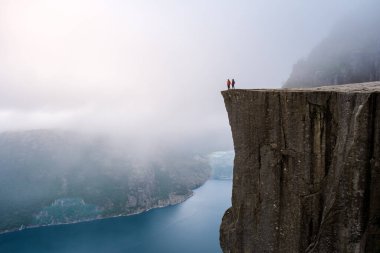 Norveç 'teki Kürsü Kayası' nın tepesinden bir manzara, iki kişi kıyıda duruyor, sisli fiyorda bakıyor. Norveç, Preikestolen