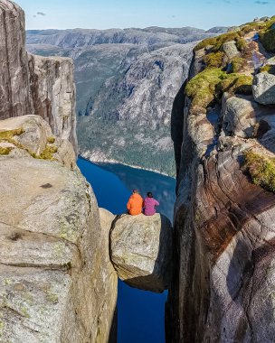 İki yürüyüşçü, Norveç 'teki iki yüksek uçurum arasındaki uçurumu birbirine bağlayan bir kaya oluşumunun üzerinde oturur ve çevresindeki manzaranın nefes kesici manzarasının tadını çıkarırlar. Kjeragbolten Norveç