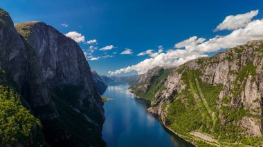 Pofuduk bulutlarla kaplı açık mavi bir gökyüzünün altında yükselen Norveç fiyordunun nefes kesici bir görüntüsü. Kjerag, Lysebotn, Lysefhjorden, Norveç