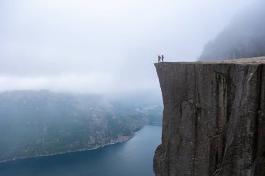 Preikestolen 'in kıyısından panoramik bir manzara, Norveç' te bir uçurum, sisli bir fiyorda tepede duran iki figürü gösteriyor. Preikestolen, Norveç 'te bir çift kadın ve erkek.
