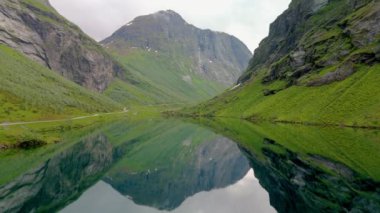 Yüksek dağlarla çevrili sakin bir gölün videosu. Dağ ve çimlerin berrak sularda yansıdığı sakin bir manzara. Norveç fiyortları Stavbergvatnet Urke