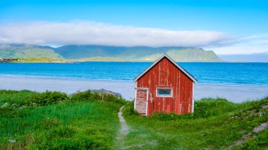 Norveç 'te, turkuaz bir denizi ve yeşil bir dağ sırası olan beyaz bir kumsal üzerinde yıpranmış kırmızı bir kulübe bulunur. Ramberg Plajı Lofoten Norveç