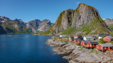 A scenic view of red cabins built on stilts along the rocky shore of a fjord in Norway. Hamnoy fishing village on Lofoten Islands, Norway with red rorbu houses clipart